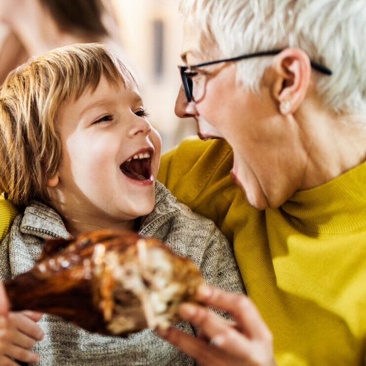 Happy boy and his grandmother having fun while about to eat turkey leg during family's lunch at home.