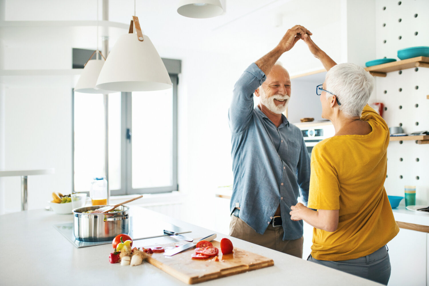 Closeup side view of a mid 60's couple having fun while cooking lunch together. They are dancing next to kitchen counter as the vegetable soup is slowly simmering.