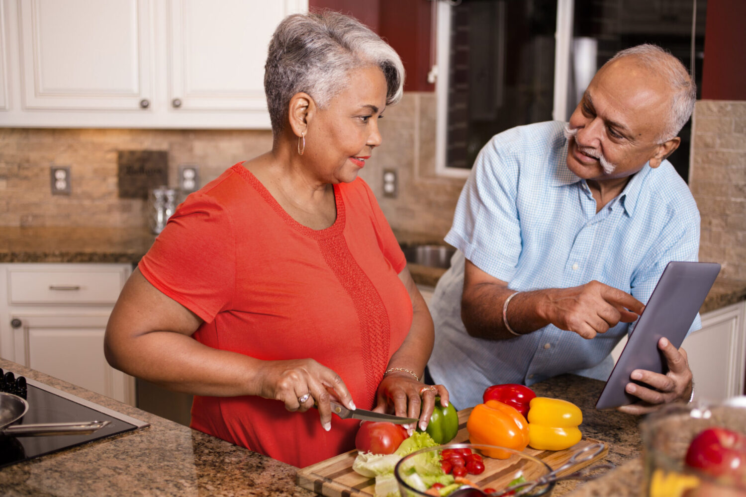 Senior adult (Indian and African descent) couple cooking dinner together at home. They are enjoying their retirement years as they work together to chop healthy vegetables.  Man shows woman recipe on digital tablet.