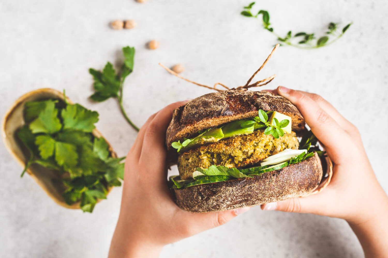 Vegan sandwich with chickpea patty, avocado, cucumber and greens in rye bread in children's hands, top view.